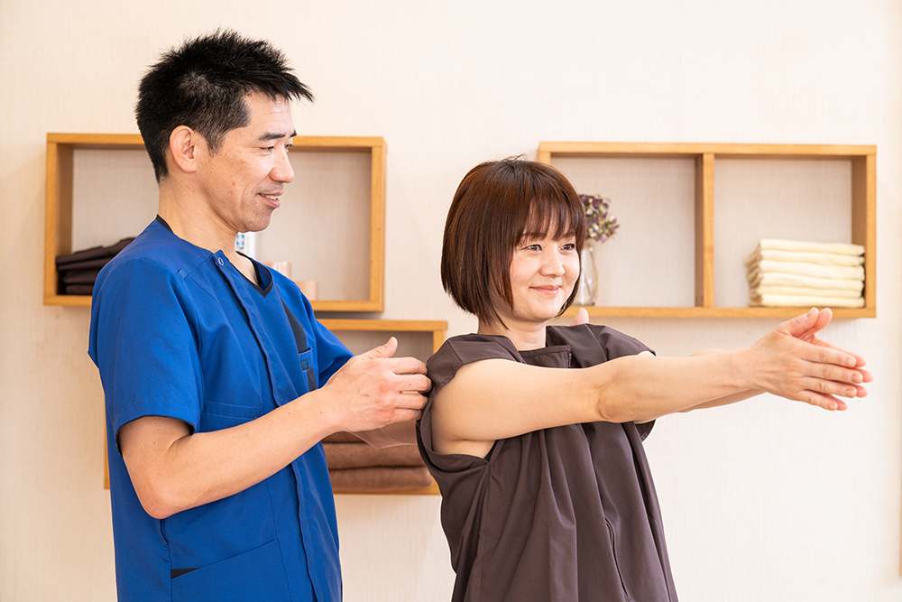 立って両腕を前に伸ばす人の肩に手を添えて、動きを確かめる施術者